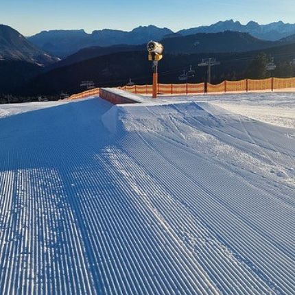 A well-groomed ski slope with orange fencing and ski lifts in a mountain setting under a clear blue sky.