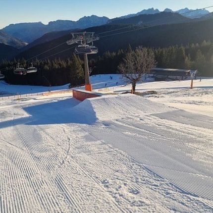 Schneebedeckte Skipiste mit Skihügel und Schlepplift, umgeben von verschneiten Bäumen und Bergen bei klarem Himmel.
