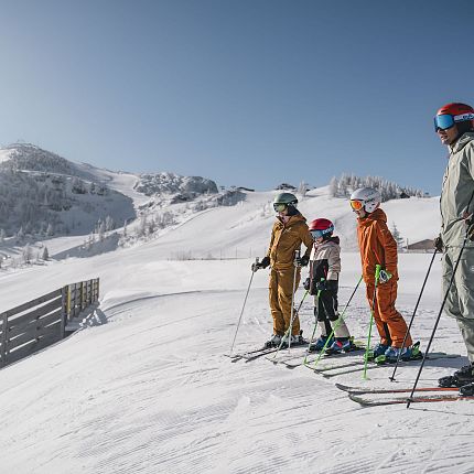 Familie auf Skiern in einem schneebedeckten Berggebiet unter klarem Himmel.
