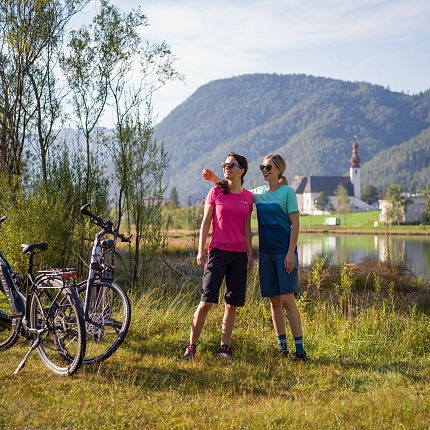 Ein Paar mit Fahrrädern vor einer Berg- und Seelandschaft, mit einer Kirche im Hintergrund.