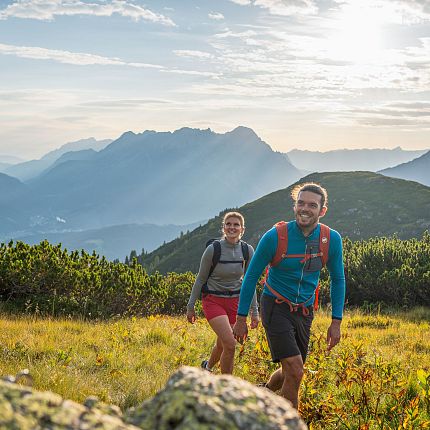 Zwei Personen beim Wandern in Berglandschaft mit Sonne im Hintergrund.