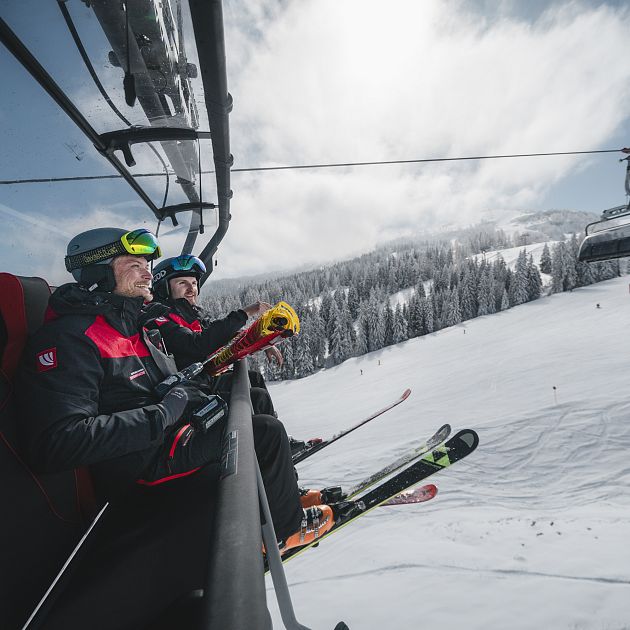 Zwei Skifahrer sitzen auf einem Sessellift in einer verschneiten Berglandschaft.