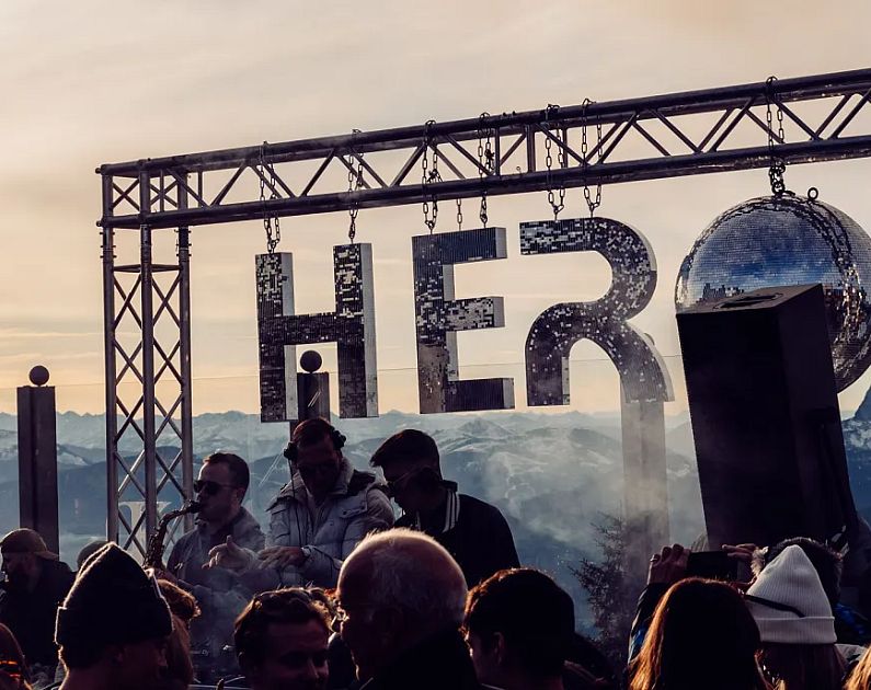 Menschenmenge bei Sonnenuntergang auf einem Berggipfel, Metallkonstruktion mit 'HERO'-Schriftzug im Hintergrund.