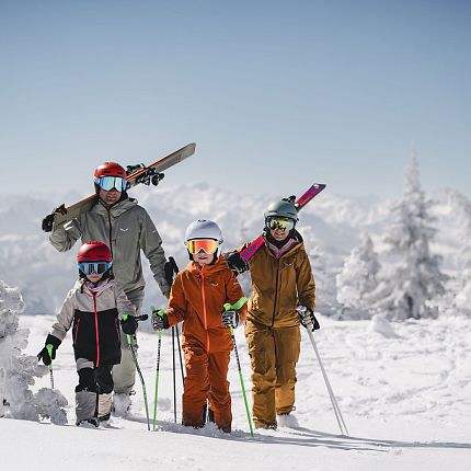 Family of four in ski gear on a snowy mountain with clear skies and snow-covered trees in the background.