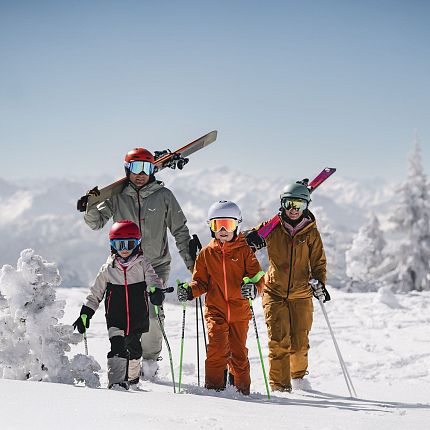 Family of four in ski gear on a snowy mountain with clear skies and snow-covered trees in the background.