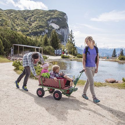 A family walks by a lake in a mountainous area. Parents pull a wagon with kids, surrounded by nature, clear skies, and distant mountains.