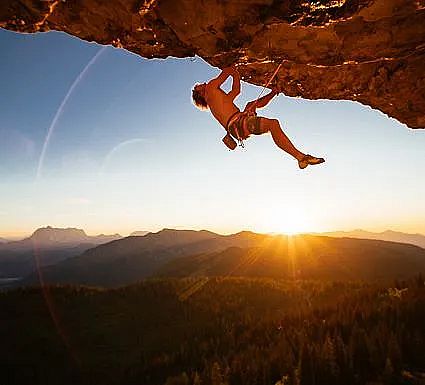A person rock climbing on an overhang with a sunset and mountains in the background.