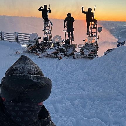Group of people with snowmobiles on snowy terrain during a sunset, orange glow in the sky, and snow covered mountains in the background.