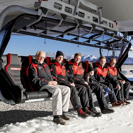 Sechs Personen sitzen auf einem Sessellift im Schnee, im Hintergrund Berge.