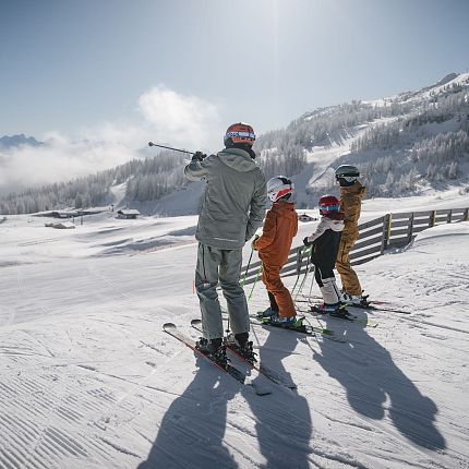 Drei Skifahrer stehen auf einer verschneiten Bergpiste mit wolkigem Himmel im Hintergrund.