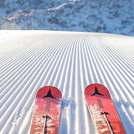 Red skis on a groomed snowy slope with mountains in the background.