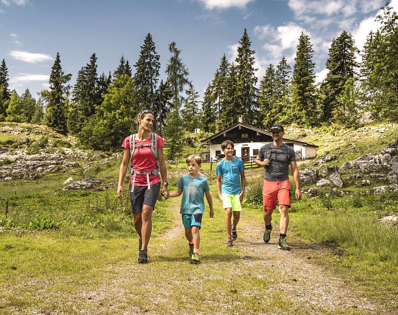 Family hiking on a trail in a scenic mountainous landscape with trees and a cabin in the background.